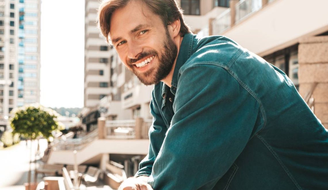 Portrait of handsome smiling stylish hipster lambersexual model.Man dressed in green shirt. Fashion male posing in the street at skyscraper background